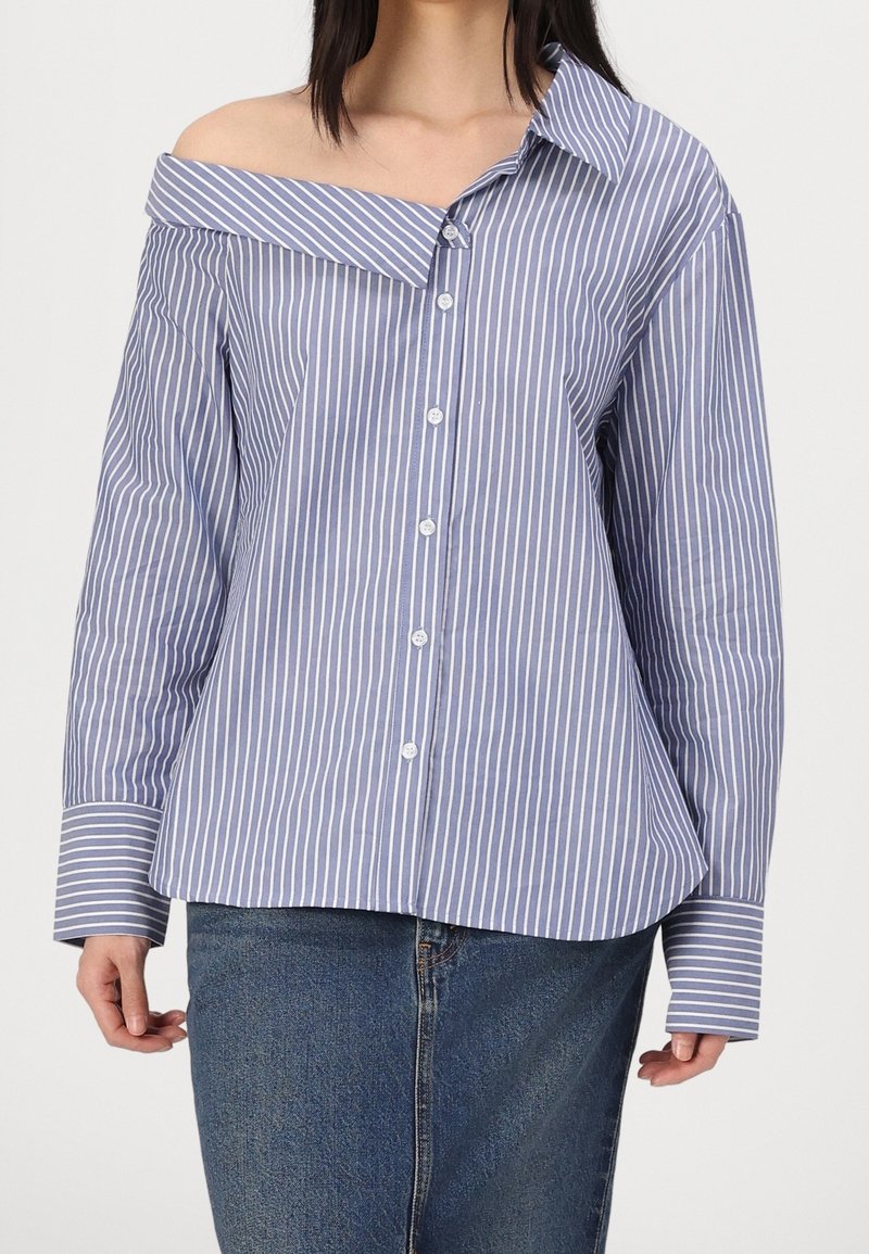 Femme portant une chemise à rayures verticales bleu et blanc, épaules dénudées, boutonnée, associée à un jean en denim bleu.