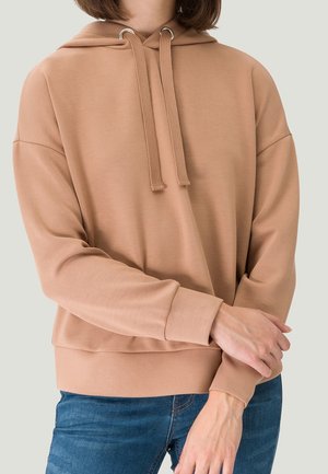 Personne portant un sweat à capuche marron clair avec des cordons et un jean bleu, bras croisés à la taille devant un fond uni.