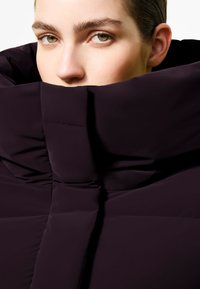 Giacca scura e voluminoso con un colletto alto e strutturato; texture morbida e tessuto liscio; vestibilità sagomata che mette in risalto un profondo colore prugna.