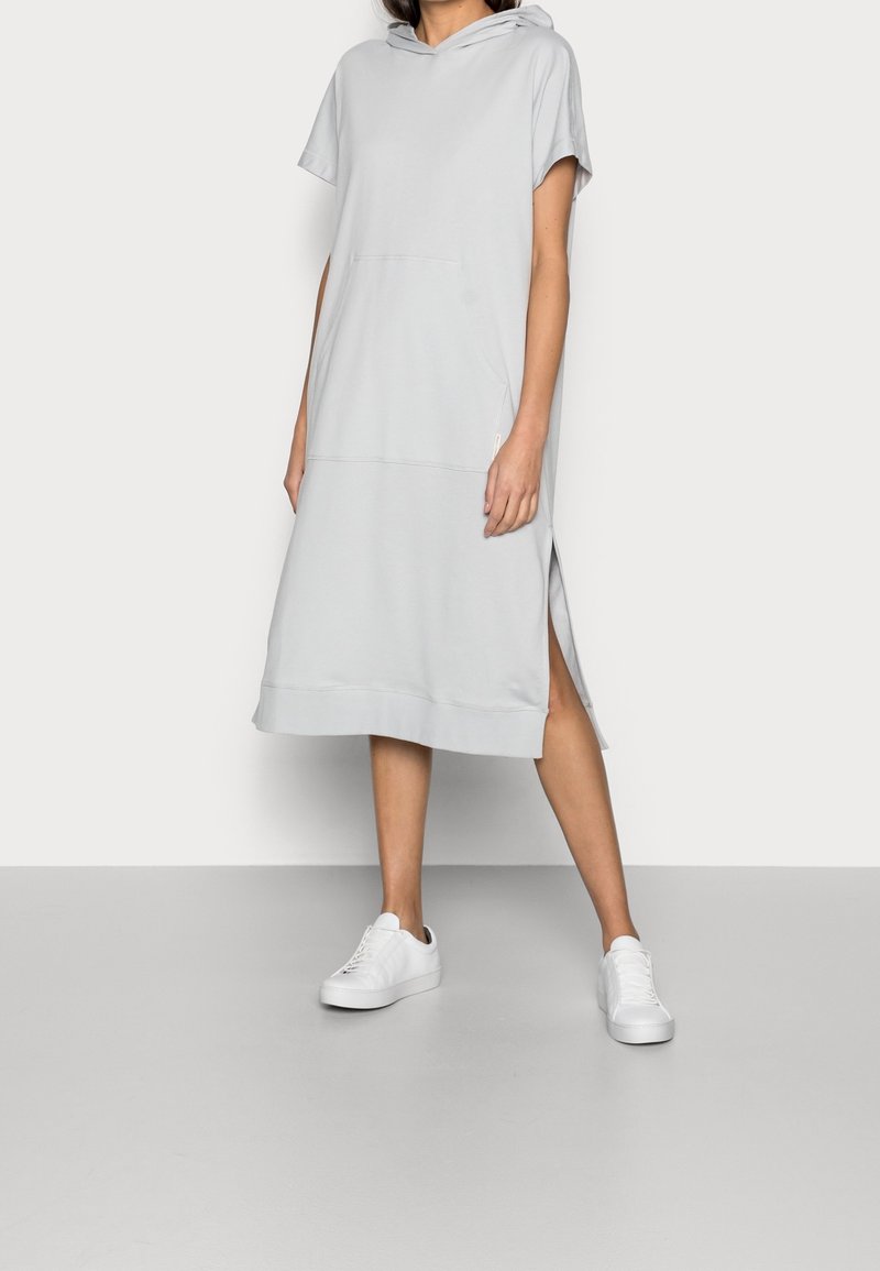 Robe à capuche gris clair avec des manches courtes, poche avant, fentes sur les côtés et ourlet côtelé, associée à des baskets blanches sur un sol neutre.