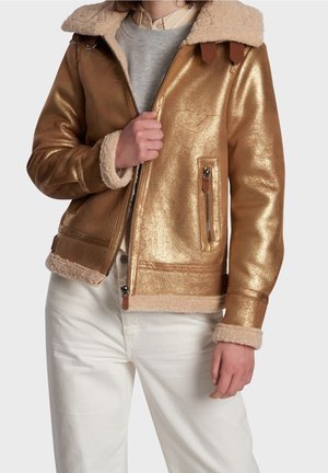 Personne portant une veste en peau retournée dorée brillante avec une doublure et des poignets en fourrure beige, un pantalon blanc et un haut superposé beige et gris, tenant la fermeture éclair de la veste.