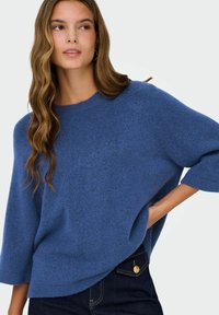 Femme aux longs cheveux ondulés portant un pull bleu oversize et un jean foncé, posant avec une main sur la hanche devant un fond uni.