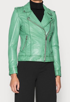 Vihreä nahkainen moottoripyörätakki hopeisilla vetoketjuilla ja neppareilla, päällä musta poolopaita ja mustat housut.