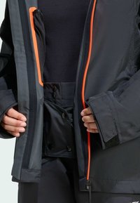 Svart vattentät jacka med orangea dragkedjedetaljer, en strukturerad innerficka och tryckknappar. Materialet verkar vara syntetiskt med en slät yta.