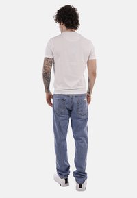 Homme aux cheveux bouclés et tatouages sur les bras, portant un t-shirt blanc, un jean bleu et des baskets blanches, debout de dos sur un fond blanc.