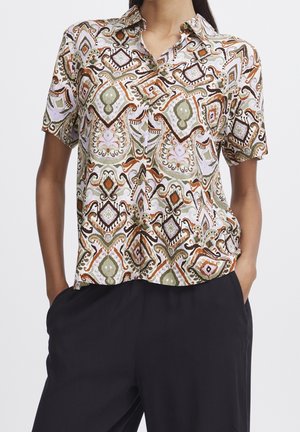 Kobieta nosi krótką koszulę na guziki z wielokolorowym geometrycznym wzorem i czarne luźne spodnie, ręce w kieszeniach, na jednolitym tle.