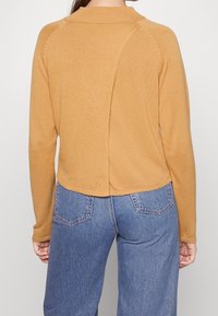 Maglione giallo senape con maniche lunghe, tessuto a coste e orlo arrotondato. Presenta un dettaglio della cucitura posteriore e un colletto alto. Abbinato a jeans blu.