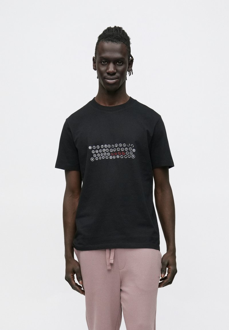 Juoda medvilninė marškinėliai su grafiniu dizainu, kuriame yra baltos ir rausvos spalvos apskritimo motyvai, apvali iškirptė, dėvimi su šviesiai rožiniais sportiniais kelnėmis.