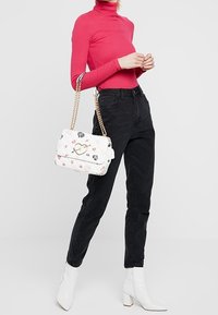 Bílá kabelka s květinovými vzory a zlatými detaily, spárovaná s růžovým topem s dlouhým rukávem a černými džíny, doplněná bílými kotníkovými botami.