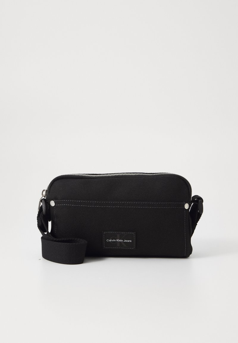 Borsa a tracolla nera in tela con chiusura a zip, tracolla regolabile e patch con logo. Presenta dettagli di cucitura a contrasto.