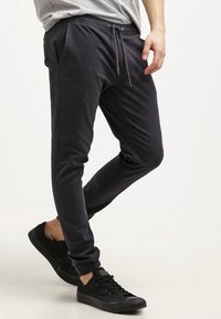 Černé teplákové kalhoty z hladké tkaniny s elastickým pasem, bočními kapsami a olemovanými kotníky, v kombinaci s černými teniskami.