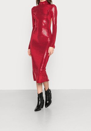 Femme portant une robe rouge à sequins à manches longues et col montant, associée à des bottines noires à boucles, debout devant un fond uni.