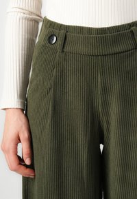 Pantalons en velours côtelé vert olive avec une texture rayée verticale, présentant un devant plat et un détail de bouton noir à la taille.