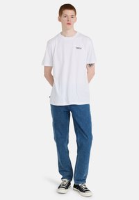 Camiseta de algodón blanca de manga corta, con cuello redondo y un pequeño logo impreso en el pecho, combinada con jeans de mezclilla azul y zapatillas negras.