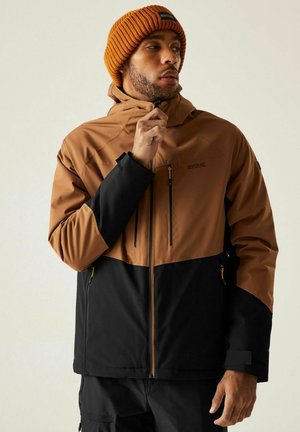 Mężczyzna w brązowo-czarnej kurtce outdoorowej i pomarańczowej dzianinowej czapce, zapinający zamek kurtki, patrzący w bok na neutralnym tle.