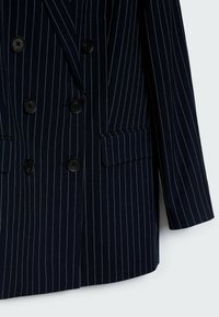 Laivastonsininen pinstripe-bleiseri, kaksirivinen malli mustilla napeilla, teksturoitua kangasta, kaksi sivutaskua ja mittatilausmainen leikkaus.