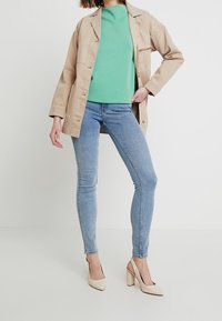 Beige oversized jacka över en mintgrön sweatshirt, kombinerad med ljusblå skinny jeans och beiga högklackade skor. Släta texturer.