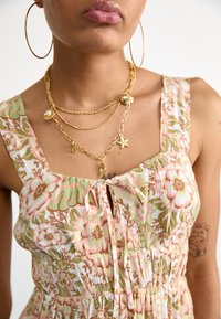 Bloemendress met roze en groene patronen, met een vierkante halslijn, verstelbare strik en zachte stof. Gouden gelaagde kettingen met bedels.