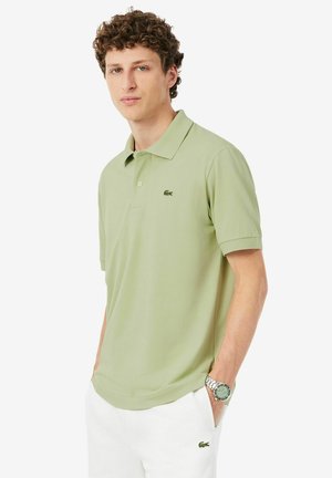Mann mit lockigem Haar, der ein hellgrünes Poloshirt und weiße Hose mit Krokodil-Logo trägt, Hände in den Taschen und eine silberne Armbanduhr.