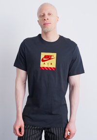 Άνδρας με αλβινισμό που φοράει μαύρη μπλούζα Nike Air και μαύρο παντελόνι με ρίγες, σταντ σε απλό ανοιχτό φόντο.