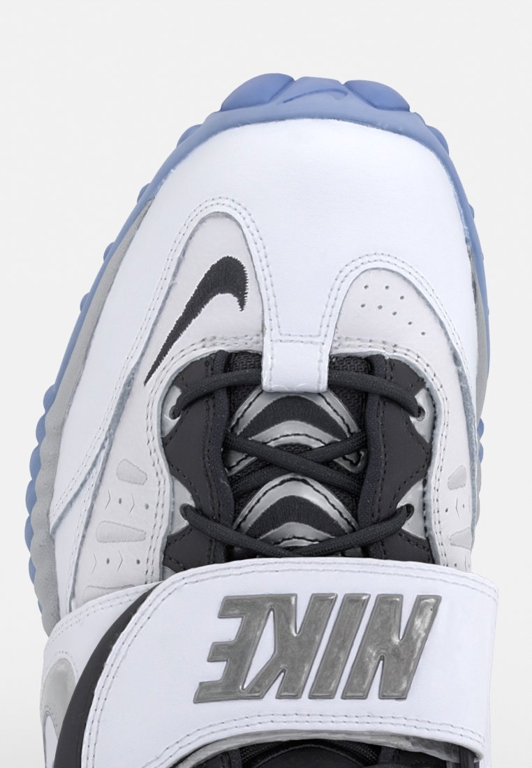 Zapatilla Nike con parte superior blanca, acentos negros y detalles grises. Presenta cordones redondos, suela de goma con toques azules y un logo prominente.