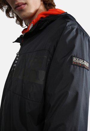 Schwarze Kapuzenjacke aus glattem Nylonmaterial mit auffälligem Logo-Print und einem Patch-Detail am Ärmel. Orangefarbenes Kapuzenfutter.