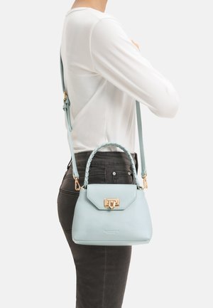 Person mit weißem Langarmshirt und dunkler Hose hält eine hellblaue Handtasche mit geflochtenem Griff und goldfarbener Verschluss an der Seite.