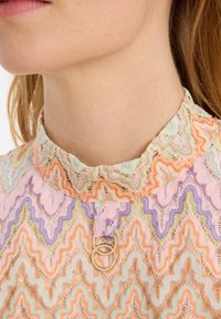Spitzenbluse mit gewelltem Kragen, die ein Zickzackmuster in Pastellfarben (Rosa, Orange, Lila) und ein metallisches Ringdetail am Hals aufweist.