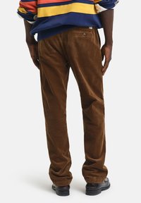Brown corduroy pants with vertical ribs and a straight-leg design, featuring back pockets and a subtle logo detail.
