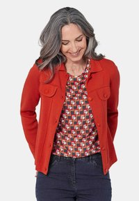 Rode gebreide jas met knoopsluitingen en twee borstzakken, gecombineerd met een top met geometrisch patroon in meerdere kleuren. Model draagt een donkere jeans.