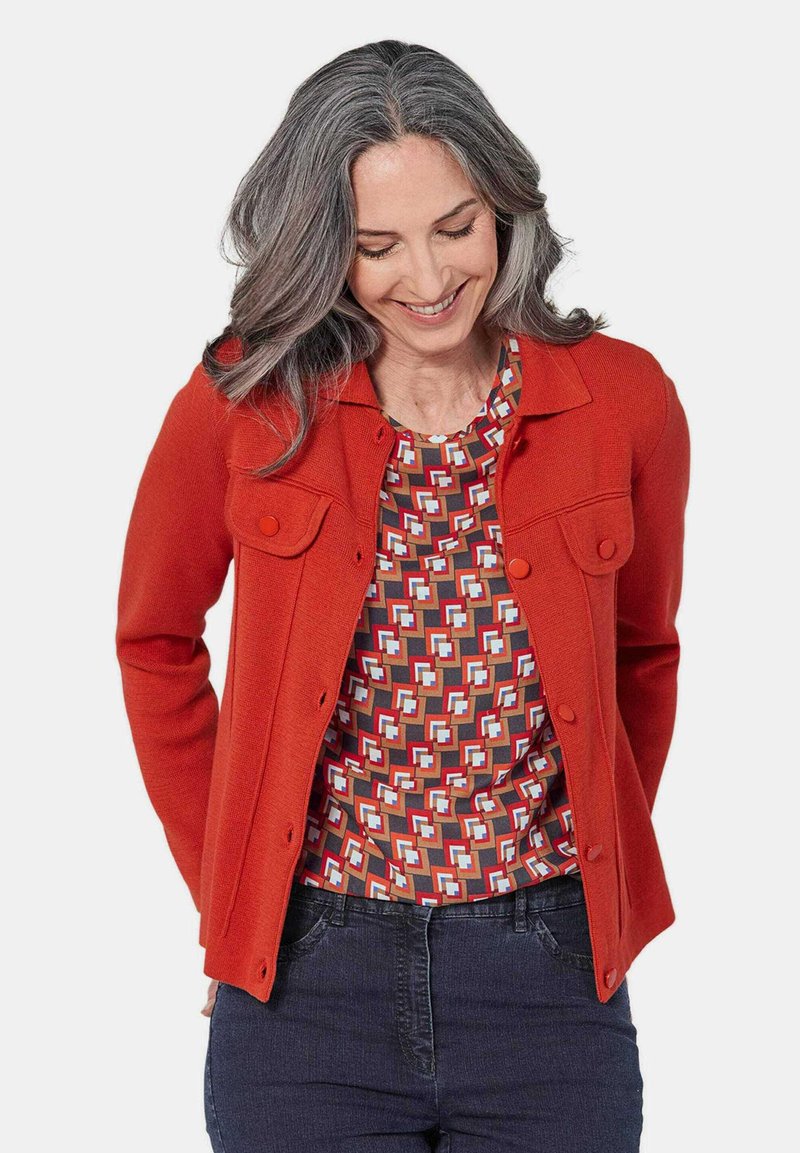 Rode gebreide jas met knoopsluitingen en twee borstzakken, gecombineerd met een top met geometrisch patroon in meerdere kleuren. Model draagt een donkere jeans.
