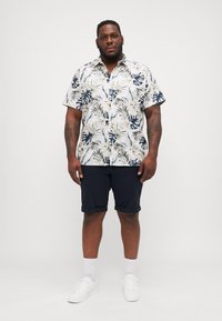 Camisa de manga corta, con botones, con un patrón de hojas tropicales en verde y azul marino sobre un fondo blanco, combinada con pantalones cortos azul marino oscurecido.