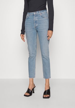 Vrouw draagt lichtblauwe rechte jeans met hoge taille, zwart top met lange mouwen en zwarte open-teen sandalen met hak, staand tegen een witte achtergrond.