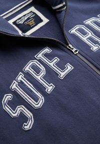 Sudadera con cremallera en color navy con texto bordado en blanco "SUPER" en la parte frontal. Presenta una textura suave y un detalle en el cuello con rayas contrastantes.