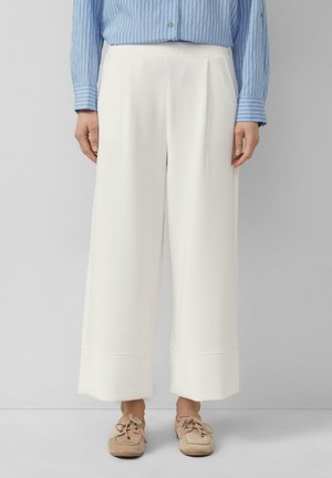 Osoba w białych szerokich, skróconych spodniach, niebieskiej koszuli w paski z długim rękawem oraz beżowych mokasynach stoi na tle jednolitego tła.