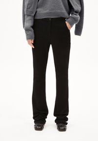 Person, der schwarze gerade geschnittene Hosen, schwarze Fransen-Schlüpfschuhe und einen grauen, flauschigen Pullover trägt, mit einer Hand in der Tasche vor einem weißen Hintergrund.