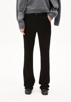 Person, der schwarze gerade geschnittene Hosen, schwarze Fransen-Schlüpfschuhe und einen grauen, flauschigen Pullover trägt, mit einer Hand in der Tasche vor einem weißen Hintergrund.