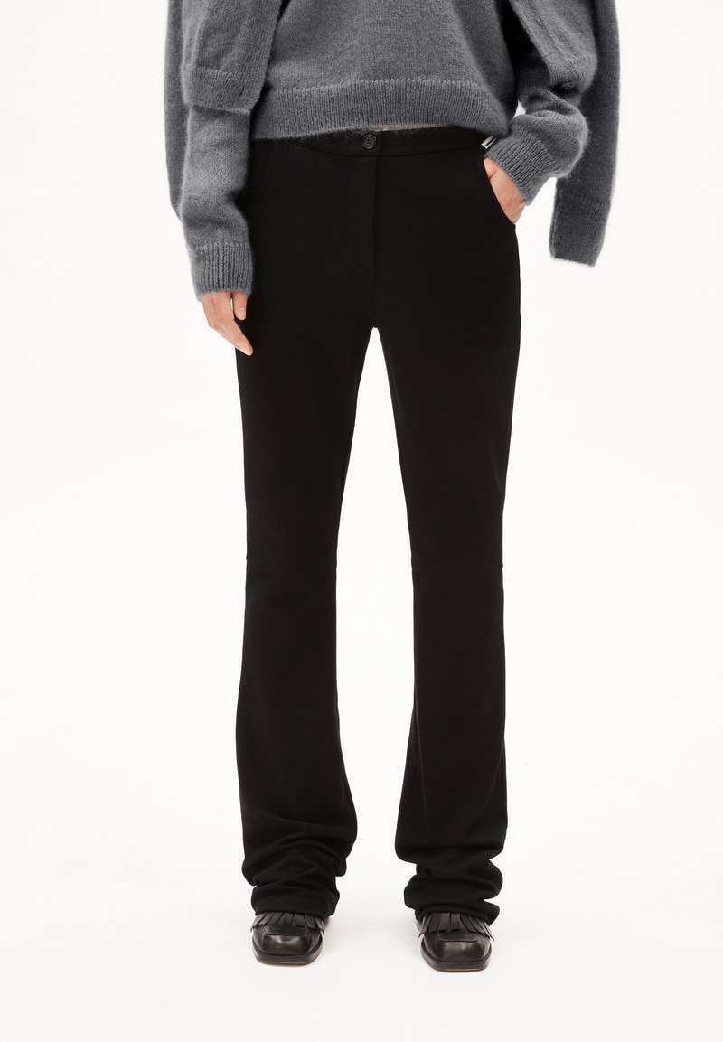 Person, der schwarze gerade geschnittene Hosen, schwarze Fransen-Schlüpfschuhe und einen grauen, flauschigen Pullover trägt, mit einer Hand in der Tasche vor einem weißen Hintergrund.