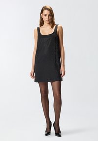 Vestido negro sin mangas con superficie texturada que presenta pequeños agujeros, con una ligera forma en A y un escote cuadrado. Llevado con medias negras transparentes.