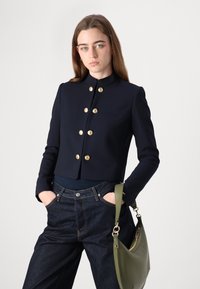 Giacca cropped navy con bottoni dorati, colletto alla cinese e una tasca sul petto. Abbinata a pantaloni in denim scuro e una borsa a tracolla verde.