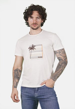 T-shirt in cotone bianco con design stampato di palme e testo "YES-ZEE", colletto rotondo e maniche corte. Indossato con jeans blu.