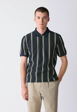 Young man standing with one hand in pocket, wearing a navy blue polo shirt with vertical beige stripes and light beige pants.