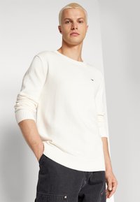 Suéter de punto color crema con cuello redondo, puños y dobladillo acanalados; presenta un pequeño logo en el pecho. Llevado con pantalones oscuros.