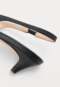 Sorte lærrem sandal med lav hæl, med en glat overflade og beige indvendig. Inkluderer en slank ankelrem med elastisk detalje.