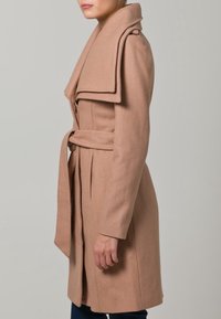 Ljust beige kappa i ullblandning med bred krage, långa ärmar, midjeband och rak skärning, med en mjuk textur och minimalistisk design.