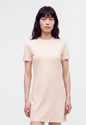 Jeune femme aux longs cheveux noirs portant une robe rose clair à manches courtes avec des bords festonnés aux manches, debout devant un fond uni.