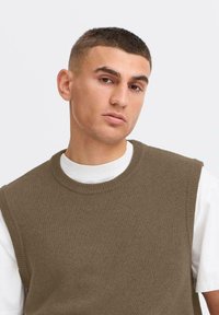 Jersey sin mangas marrón sobre una camiseta blanca. Presenta un cuello redondo y bordes acanalados, con una textura suave.