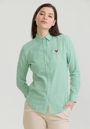 Femme portant une chemise à rayures vertes et blanches avec boutons et un patch cœur rouge sur la poitrine, pantalon beige, debout, main dans la poche.