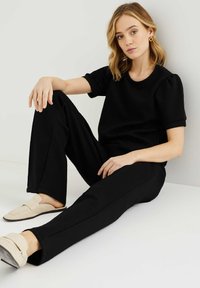 Zwarte top met korte mouwen en ronde hals, gecombineerd met zwarte rechte broeken. Beige instap schoenen maken de outfit af. Gladde stoftextuur.