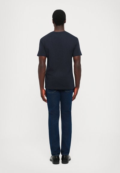 Man standing facing away wearing a dark navy t-shirt, blue jeans, and black shoes against a plain light background.